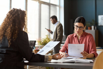 Caucasian young adult woman analyzing financial documents with another woman in office setting, Black man standing in background using smartphone near window
