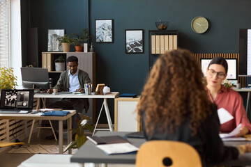 Caucasian young adult women and Black young adult man working at desks in modern office, woman with curly hair sitting in foreground, man using computer in background, documents on desks