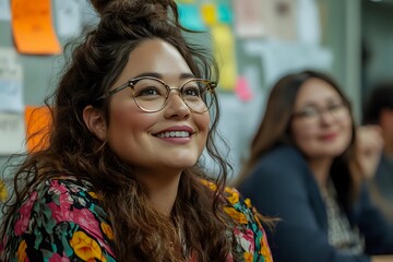 Young Hispanic woman with glasses smiling in modern office environment with colorful sticky notes on wall behind her and colleagues in background.