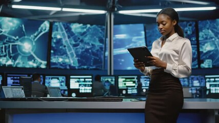 The woman is focused on her work, reviewing and assessing information on a touchscreen tablet. She appears to be in a control room with multiple screens displaying various graphics and data.