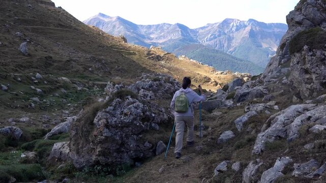 Active woman with a backpack and trekking poles descending a narrow, steep path during a hike in a beautiful mountain range in summer