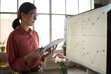Caucasian woman analyzing data charts on large monitor while holding clipboard and pen, standing in modern office environment with window background and flowers visible