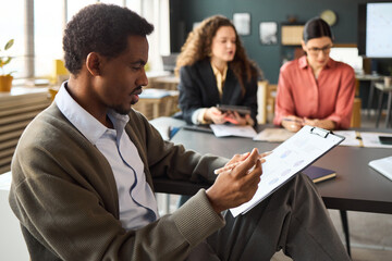 Young adult Black man analyzing business charts on clipboard while sitting at desk, two young adult Caucasian women collaborating in background, modern office setting, teamwork concept