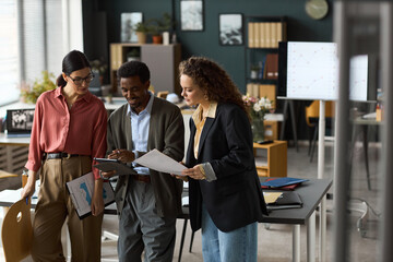 Young adult Caucasian women, young adult Black man collaborating in modern office, discussing documents and digital tablet, standing near desk with charts