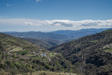 The Palancon ravine in Bayarcal, Almer&iacute;a, Spain, features rugged cliffs, terraced slopes, and lush greenery under a bright blue sky with drifting clouds. A stunning natural and rural landscape
