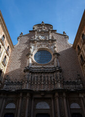 Facade of Montserrat Abbey, a Benedictine monastery in Catalonia, Spain. The Gothic and Renaissance architecture features a rose window, intricate carvings, and statues, creating a majestic scene