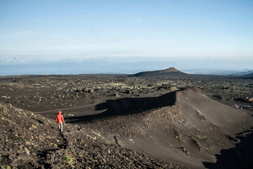 Black lava field with rugged textures and low hills, dotted with patches of green grass, as a lone person in a bright red jacket walks across the volcanic landscape.
