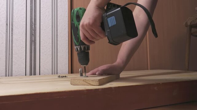 Close-up of hands using a cordless drill to fasten a metal bracket to a wooden board on a workbench. Indoor DIY woodworking and repair scene.