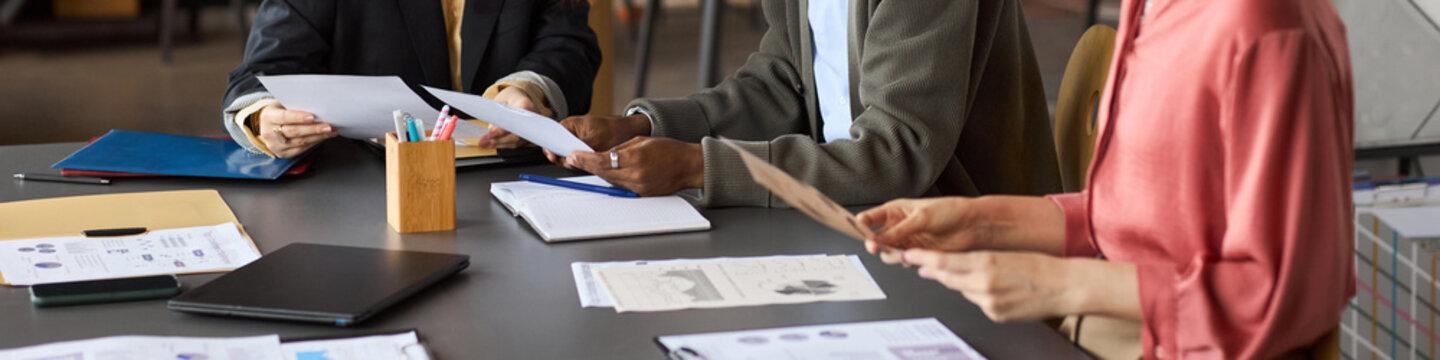 Diverse group of professionals collaborating at table, reviewing business documents and analyzing printed charts, hands of Caucasian women and Black man visible, header