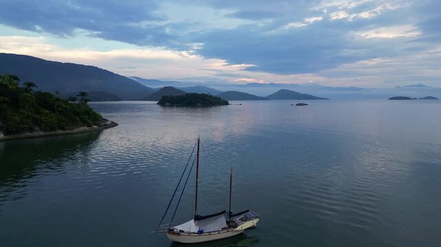 Paraty, Brazil: Aerial forward drone footage of boat anchored in Paraty, an old town in state of Rio de Janeiro, Brazil during cloudy sunset with the Costa Verde coast in the background .