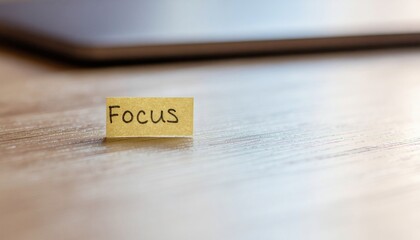 Close-up of a small yellow sticky note with the word 'Focus' written on it, placed on a light-colored wooden surface with a blurred laptop in the background, conveying a sense of motivation