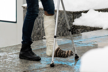 A person stands on a wet urban pavement with one leg in a white plaster cast wearing a patterned sock and black boot, using aluminum crutches beside a snow-covered curb.