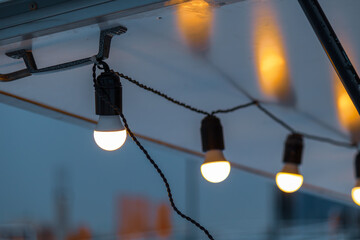 A row of glowing hanging bulbs is suspended from twisted cables beneath a metal canopy at dusk, casting warm ambient light against a softly blurred urban skyline.