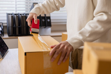 Small business warehouse scene of worker apply shipping label on parcel and taping cardboard box, organizing delivery tasks, order preparation and maintaining efficient packing workflow, multitasking