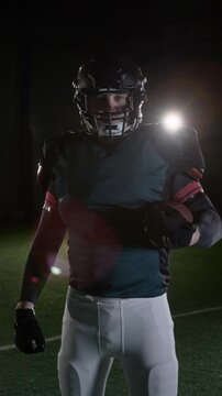 Medium vertical portrait of focused American football halfback in black jersey, protective helmet, white pants, holding ball underarm, standing at stadium, ready for match, looking at camera