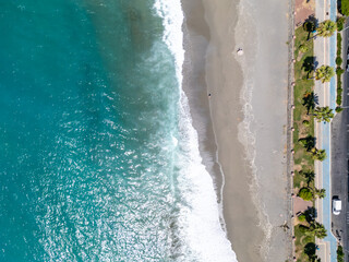 Vertical Split Aerial View of Turquoise Ocean Waves Crashing onto a Wide Sandy Beach Bordered by a Palm-Lined Coastal Road