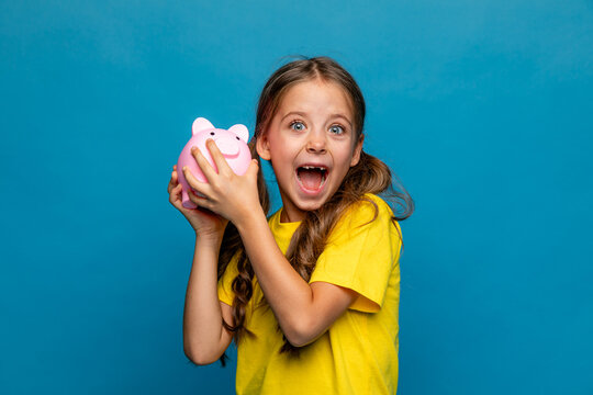 Funny little girl in yellow t-shirt holding pink piggy bank money box and looking at camera on blue background. Financial concept of children pocket money. Save money for education and family expenses