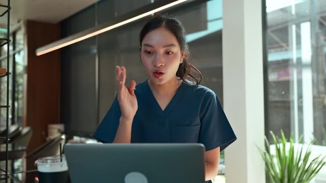 Professional female medical doctor wearing scrubs providing remote consultation through a video conference call on her laptop in an office