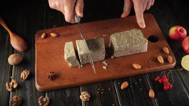Hands are cutting a block of nut-based dessert or halva on a wooden board. Nearby, there are different nuts, fresh fruit, and a small spoon. The setting is a kitchen with dark wooden surfaces.