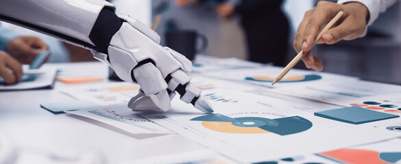 A robotic hand collaborates with human hands to analyze data charts and graphs in a modern office, illustrating the blend of technology and teamwork in business.Noogenesis