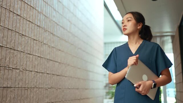 Young Asian doctor or nurse in scrubs walking confidently through a modern clinic corridor holding a laptop, ready for her next task