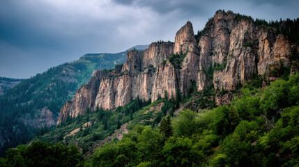 Majestic rock cliffs rise above lush green forest on a mountain slope under a dramatic sky in a stunning natural landscape of the American west region.