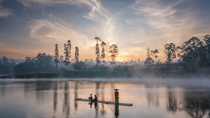 People fishing on a calm lake during sunrise with soft morning mist. Reflections on the water and silhouettes create a peaceful rural lifestyle scene in a natural outdoor environment.