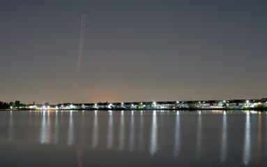 A long exposure shot showing a light trail in the sky from an airplane, and also lights and reflection on water from private houses by a tranquil lake.
