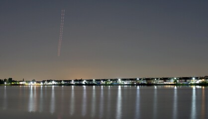 A long exposure shot showing a light trail in the sky from an airplane, and also lights and reflection on water from private houses by a tranquil lake.