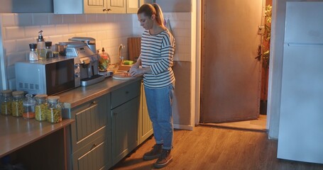 A young woman in casual clothes slices carrots on a wooden cutting board in a bright, modern...