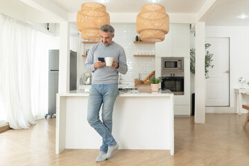 A middle-aged man enjoys a quiet moment at home, leaning on a kitchen island while using his smartphone and holding coffee. The clean interior highlights modern living and relaxed daily habits.