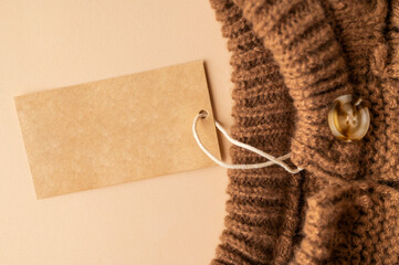 Paper cardboard tag on brown knitted wool sweater, pullover or cardigan with buttons on beige background. Children's clothing mockup, blank label for logo application. Stylish clothes fashion, cozy