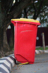 Red outdoor trash bin with a yellow lid placed on a roadside curb in an urban environment