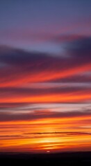 Fototapeta premium A vibrant sunset sky is painted with horizontal bands of fiery orange, red, purple, and blue hues. The bottom silhouette hints at a dark landscape