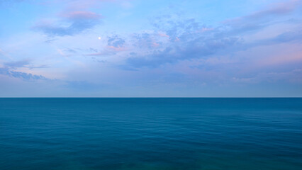 Obraz premium Sunset over sea horizon. sky blue, sun setting, moon among clouds. water calm, sky is clear.
