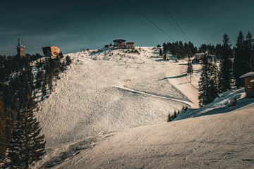 Sunlit snowy ski slope marked with tracks, featuring mountain ridge buildings, evergreen trees, and overhead lift cables against a deep sky.