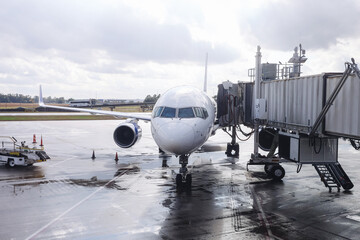 Passenger airplane parked at airport gate. Jet bridge connected to aircraft. Wet runway after rain. Commercial aviation transport scene. Travel industry infrastructure. Modern airport operations.