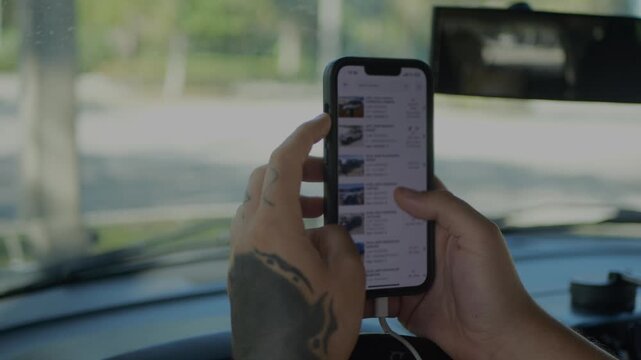 Tattooed man seated in a modern car using a smartphone, scrolling through online vehicle listings and browsing cars on a mobile car auction or dealership app.
