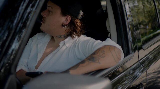 Man with tattoos sitting in car with window open, leaning on door and holding smartphone, thoughtful expression while waiting in parked vehicle, concept of driver or road trip break