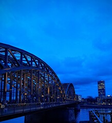 Fototapeta premium Evening landscape, city across the Rhine River in Cologne, Germany, December 19, 2025. Evening city on the riverbank. Reflection in the water. Night blue sky. 