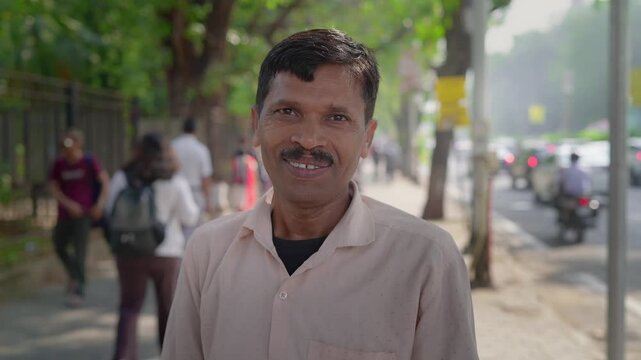 A middle-aged Indian Asian happy middle aged man or male standing outside in the daylight by a busy city road on a sidewalk, looking at the camera and smiling, with pedestrians walking in background