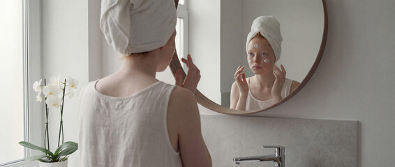 Red-haired woman applying face cream in front of round bathroom mirror with white towel wrap on head in bright minimalist space, daily morning skincare routine