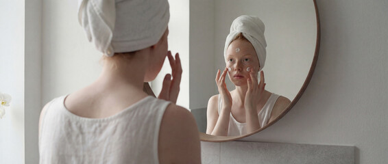 Woman with towel on head applying skincare cream in bathroom mirror after shower, self care routine in bright minimalist interior