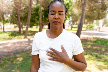 African american woman standing in a park, closing eyes, placing hand on chest, focusing on mindful breathing and meditating for balance, peace, and serenity