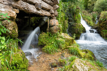 Water under pressure hits old water mill wheel, water bringed from nearby creek through metal pipe with nozzle at end, everything is covered with moss and wood is dilapidated