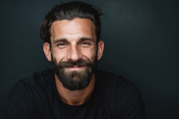 Fototapeta premium Cheerful brunette man with a beard smiles brightly in a casual black shirt against a dark background, showcasing a warm and friendly demeanor