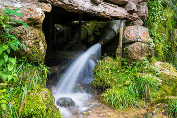 Water under pressure hits old water mill wheel, water bringed from nearby creek through metal pipe with nozzle at end, everything is covered with moss and wood is dilapidated