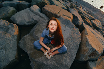 Fototapeta premium young woman sitting on rocks near water wearing denim overalls with long red hair and relaxed expression at sunset outdoors nature scene
