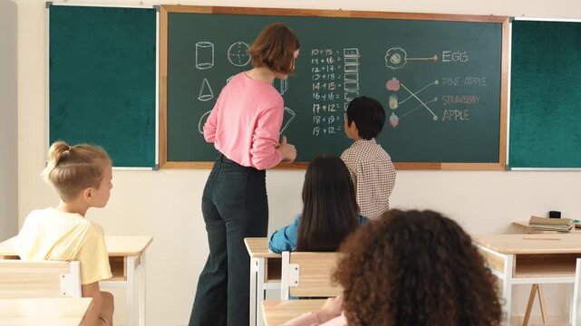 Asian smart cute boy writing answer while standing blackboard at classroom. Young beautiful teacher looking at student and learning about math lesson in elementary school. Education concept. Pedagogy.