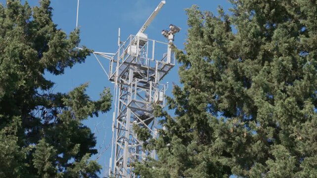 a metal communications tower featuring a white radar antenna and surveillance equipment rising above green pine trees against a blue sky.